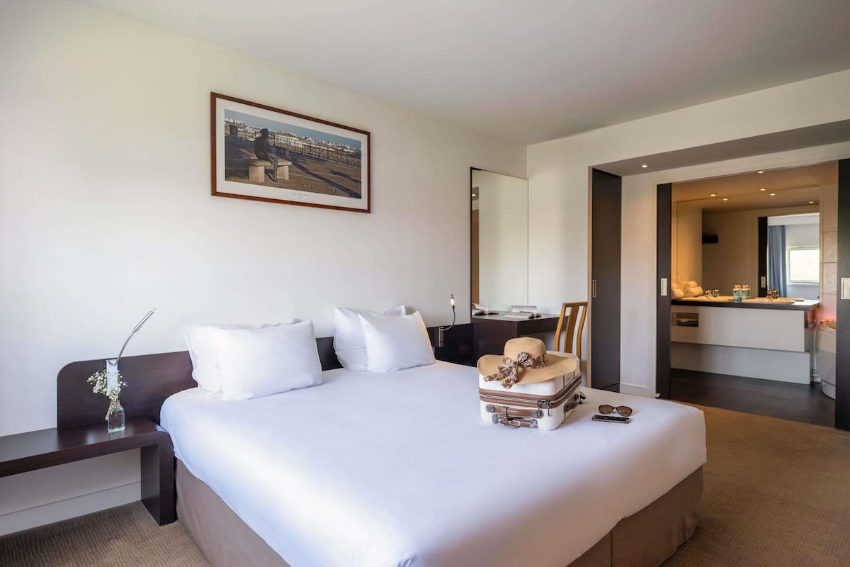 A hotel room with a large bed, a desk, a chair, a bathroom with a mirror, and a painting on the wall.