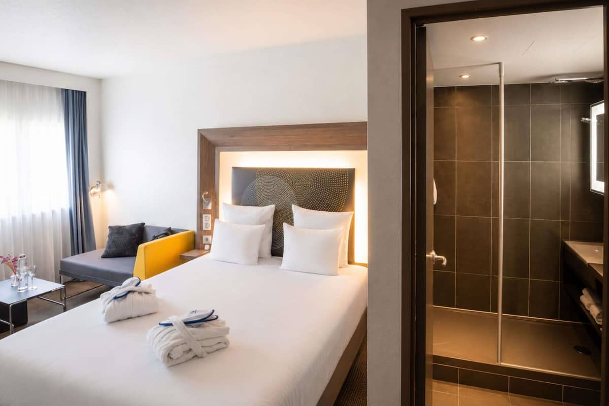 A hotel room with a bed, a desk, a chair, a shower area, and a view of the outside.