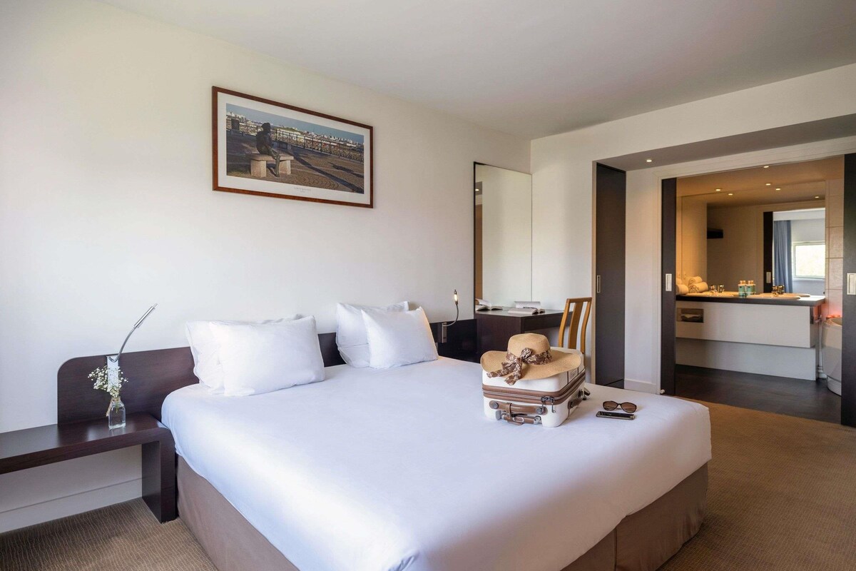 Une chambre d’hôtel avec un grand lit, un bureau, une chaise, une salle de bain avec un miroir et un tableau au mur.
