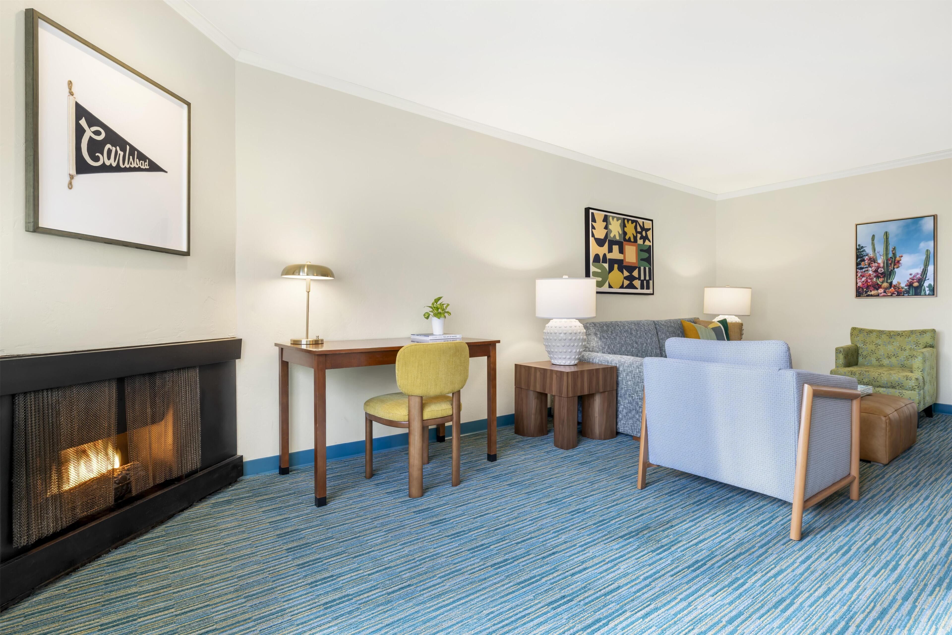 Ein Hotelzimmer mit Kamin, Schreibtisch mit Lampe, Sofa, Sessel und Kunstwerken an den Wänden.