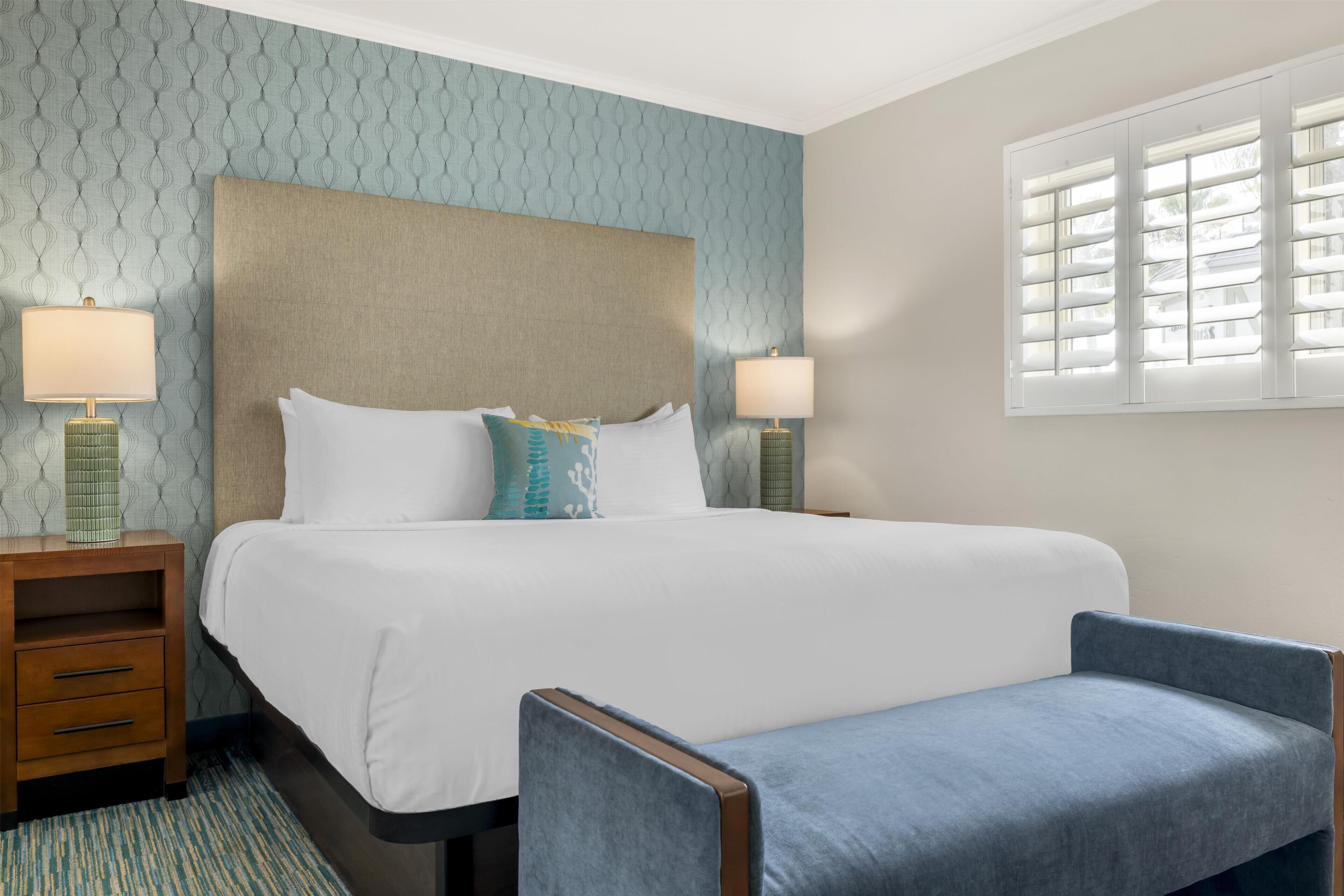 A hotel room with a large bed, two bedside tables with lamps, a blue armchair, and a window with blinds.
