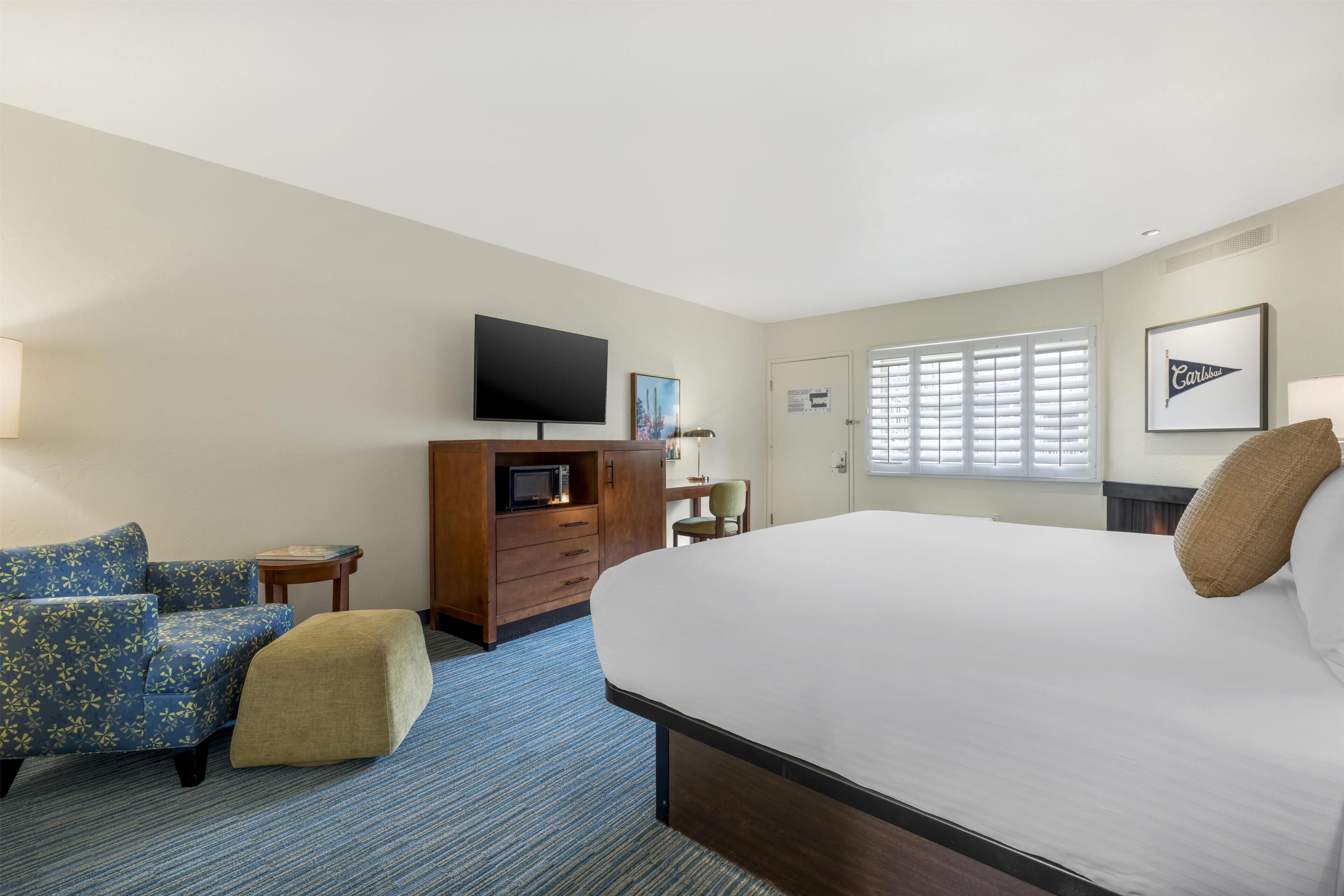 A hotel room with a large bed, a TV, a desk, a chair, and a window with blinds.
