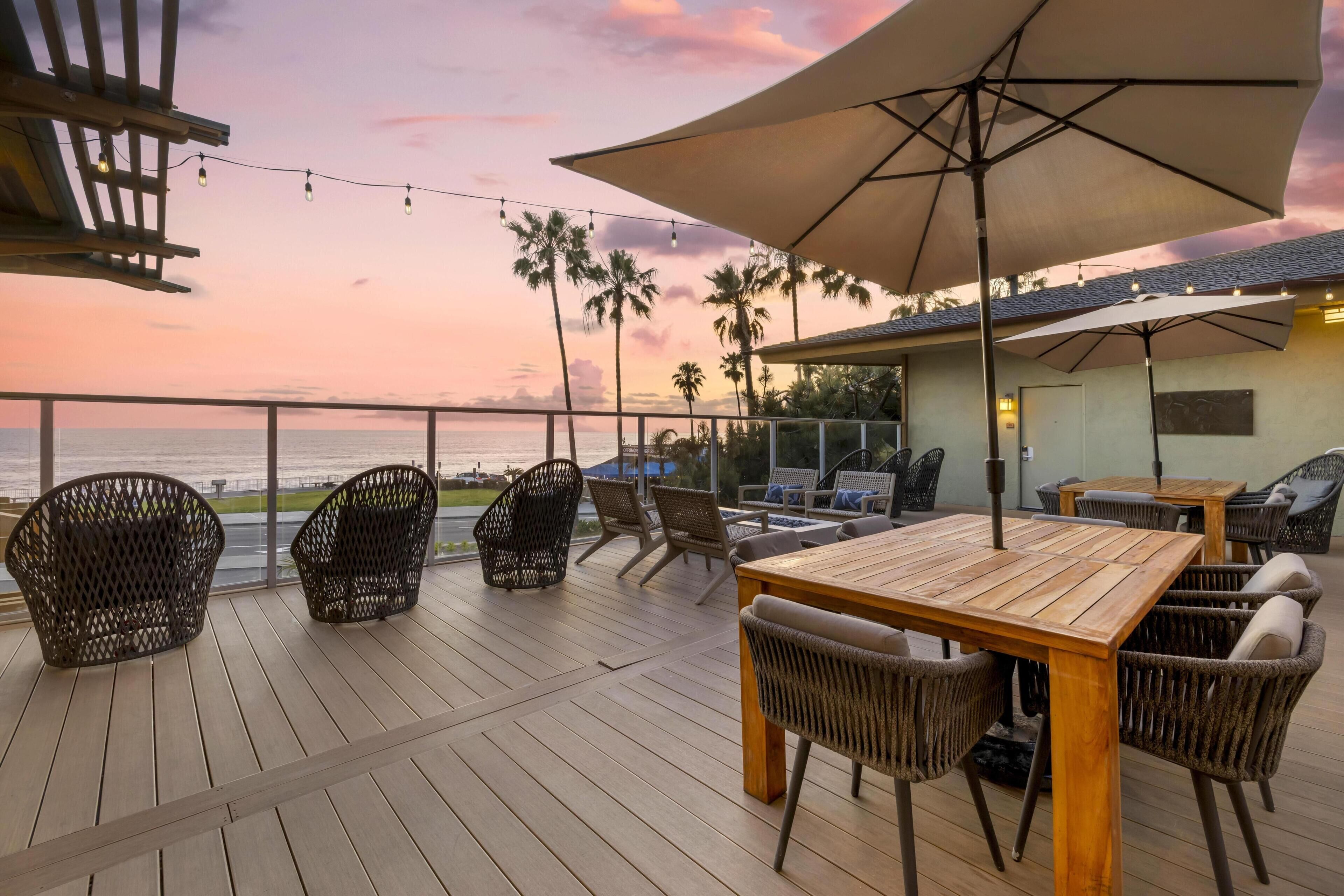Un terrazzo in legno con sedie in vimini e un tavolo, con vista sull'oceano al tramonto.