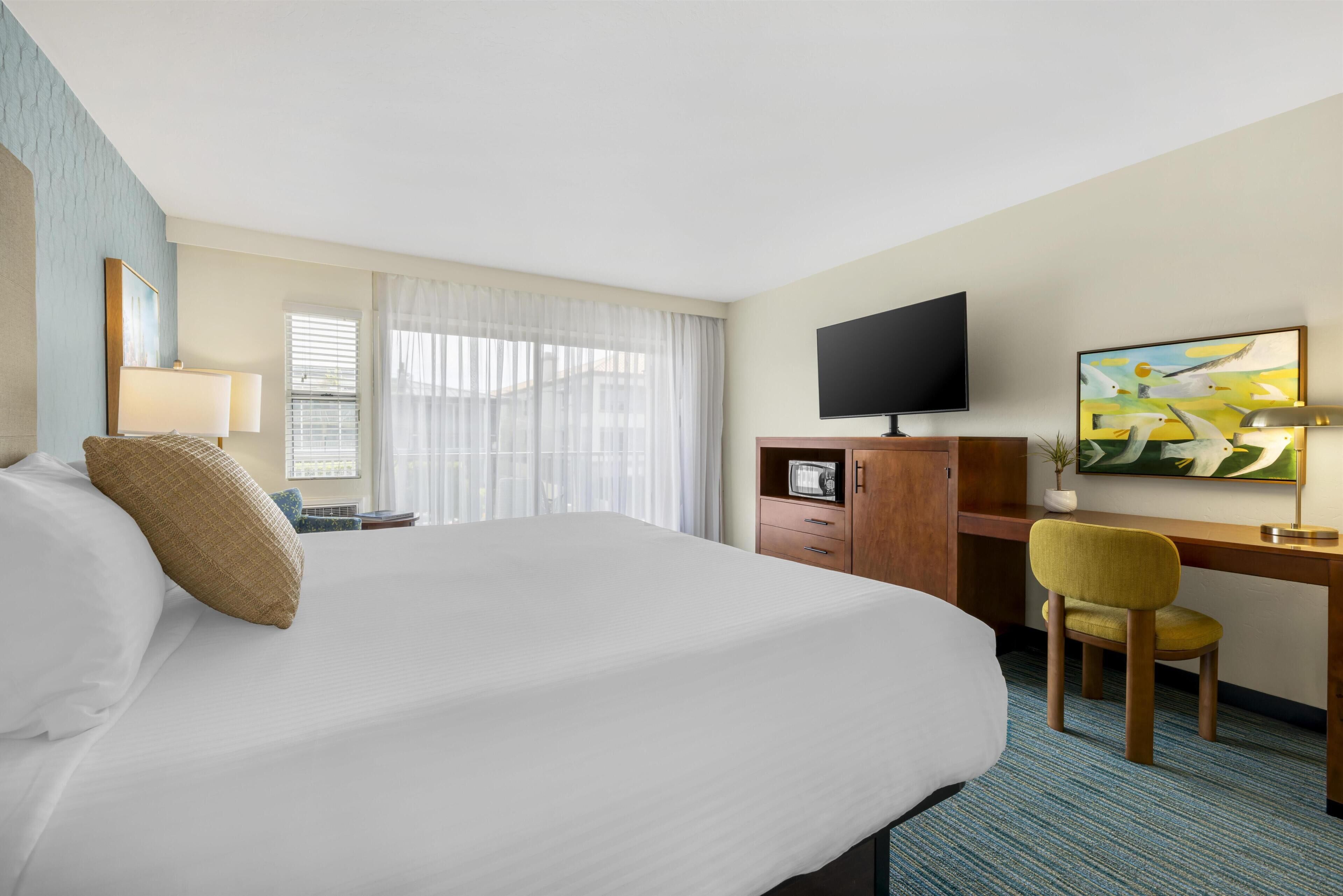 A hotel room with a bed, a desk, a chair, a television, and a painting on the wall.