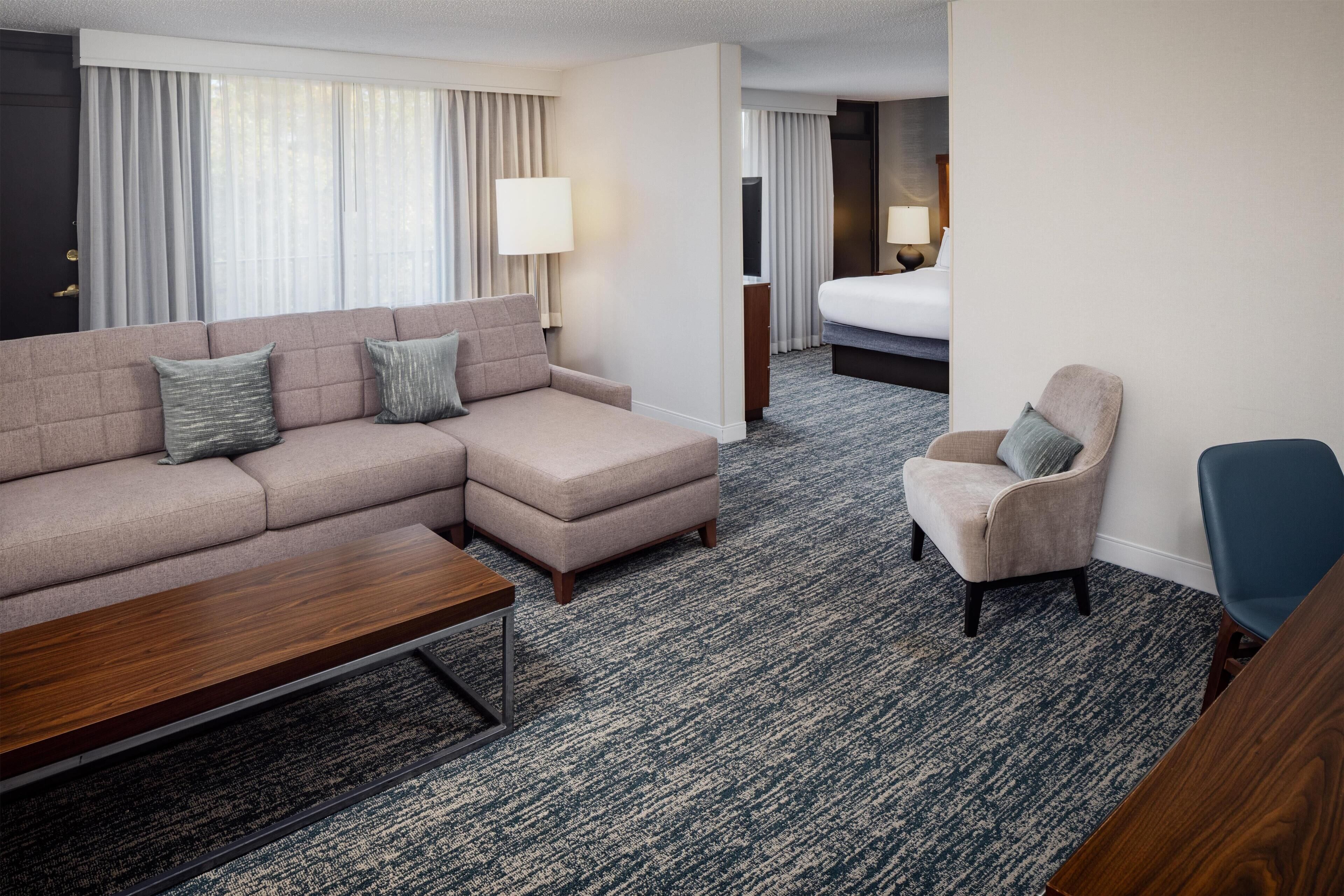 A hotel room with a sectional sofa, a single armchair, a wooden coffee table, and a bed with a nightstand.