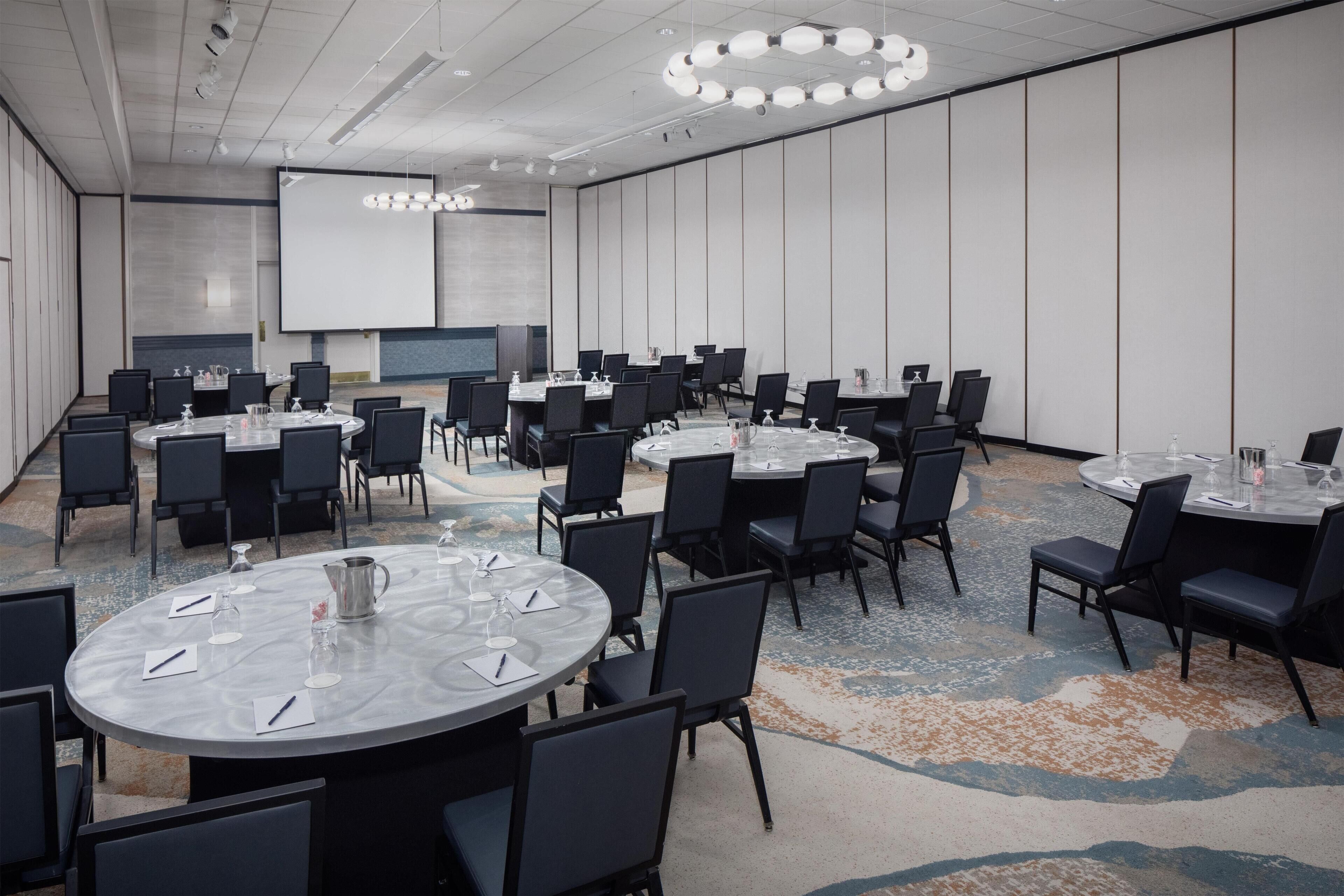 Un salón de conferencias con varias mesas redondas, sillas negras y una pantalla grande.