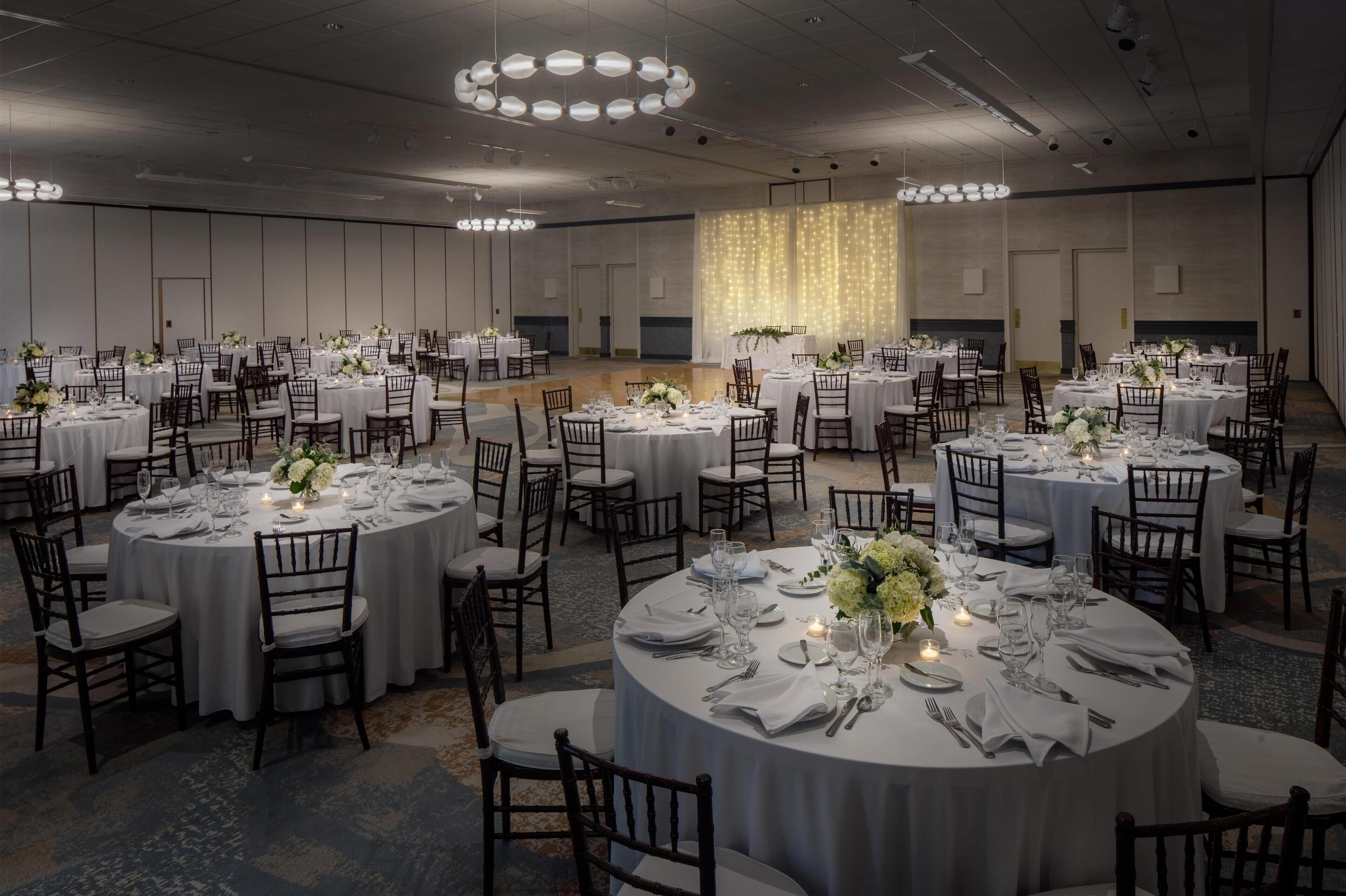 A banquet hall with round tables set for a formal event, featuring white tablecloths, floral centerpieces, and candlelight.