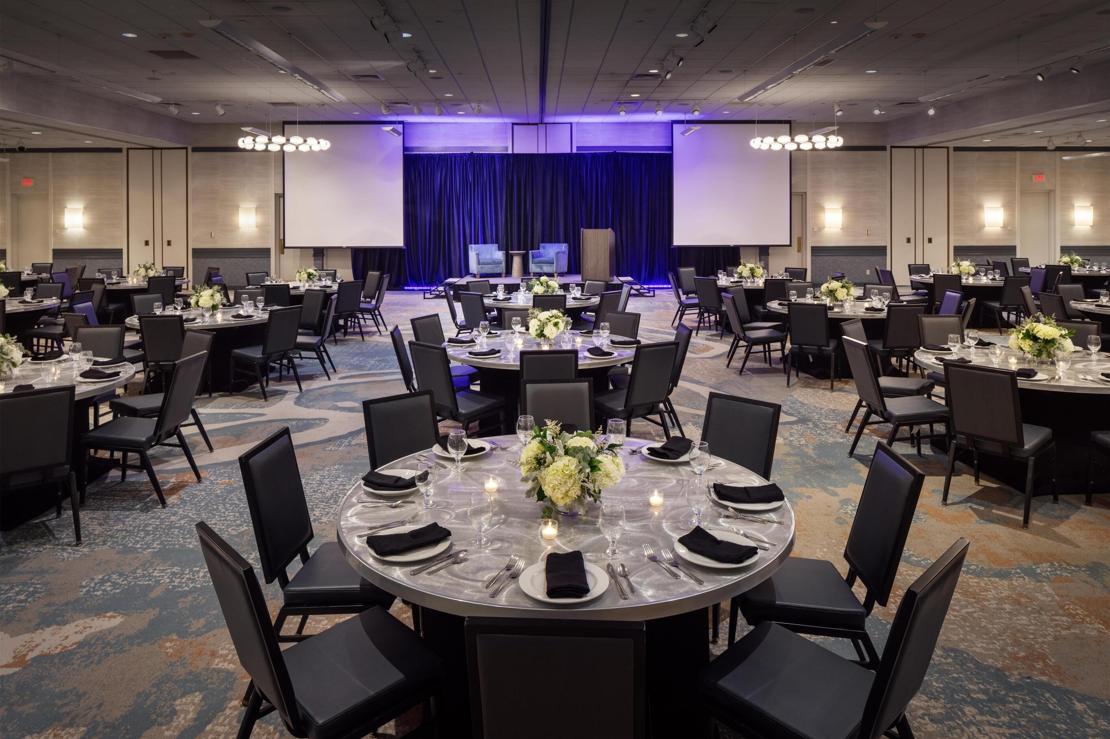 Un salón de conferencias con varias mesas redondas, cada una con sillas, platos, cubiertos y centros de mesa florales.