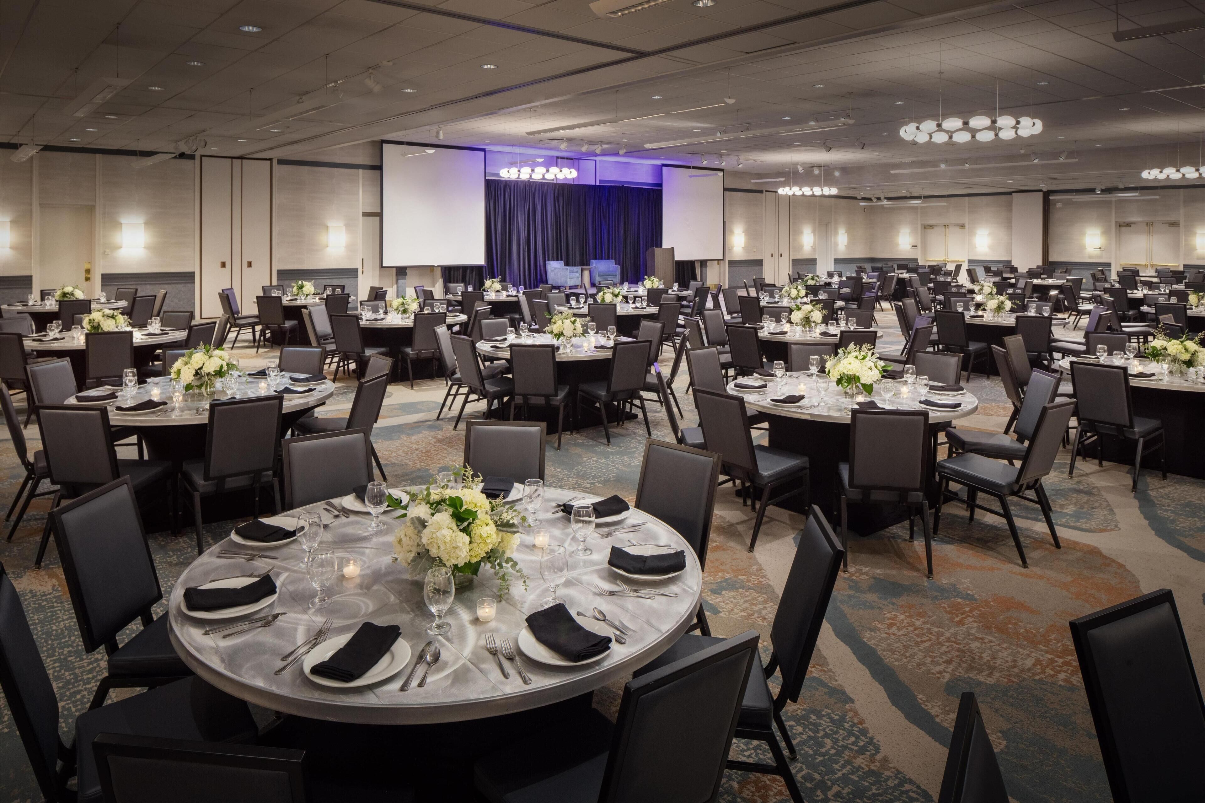 A large conference room with multiple round tables, each set with plates, cutlery, and floral centerpieces.