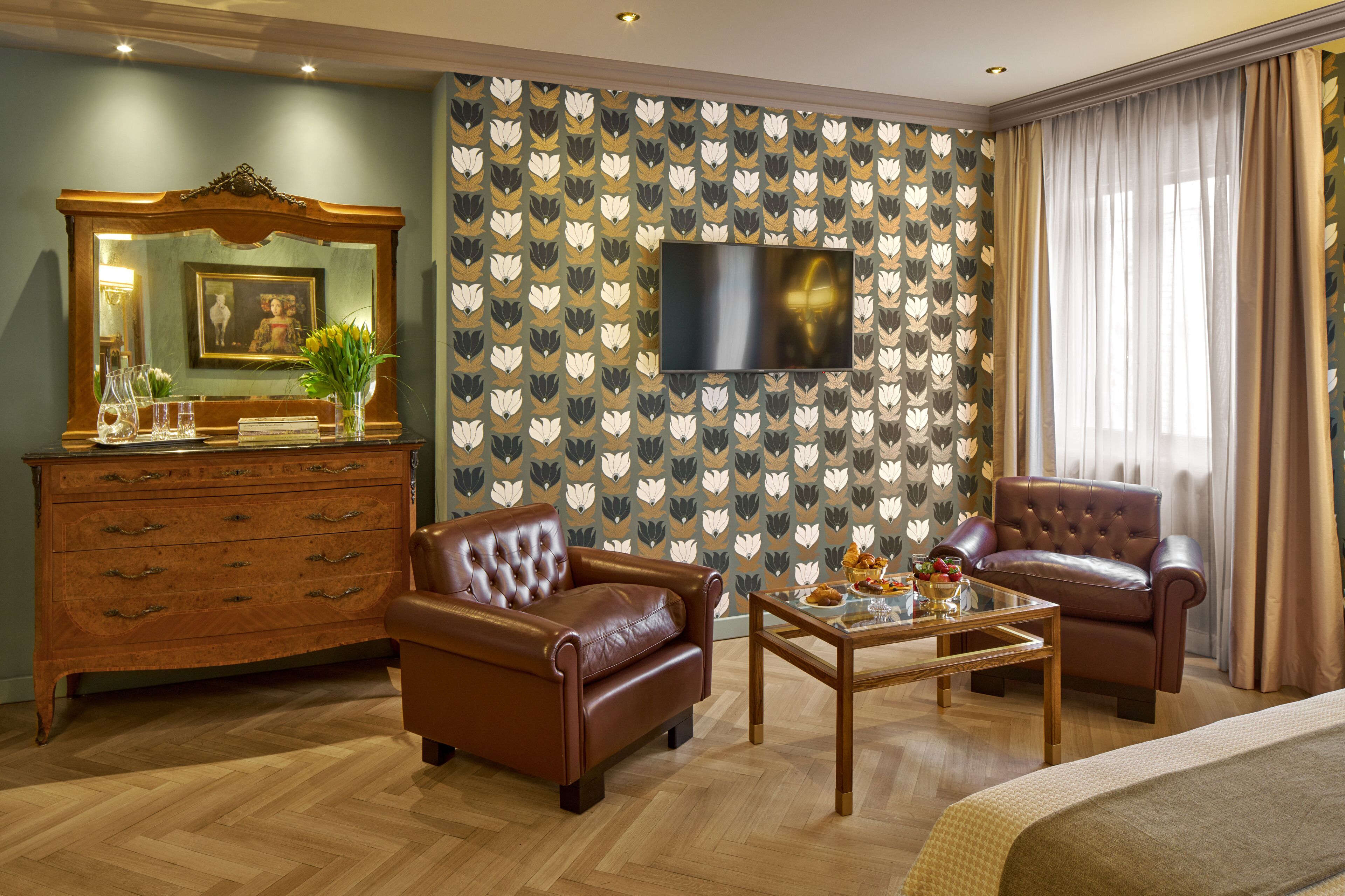 A hotel room with a wooden dresser, two leather armchairs, a glass coffee table, a flat-screen TV mounted on the wall, and a patterned wallpaper.