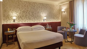 A hotel room with a large bed, two bedside tables, a round table with a chair, and a decorative vase with flowers.