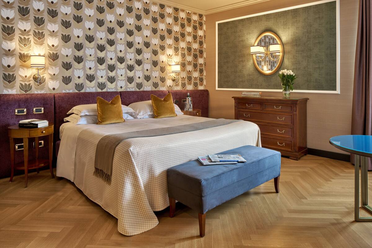 Une chambre d’hôtel avec un grand lit, une table de chevet, une commode, une table ronde et un mur orné d’un motif floral.