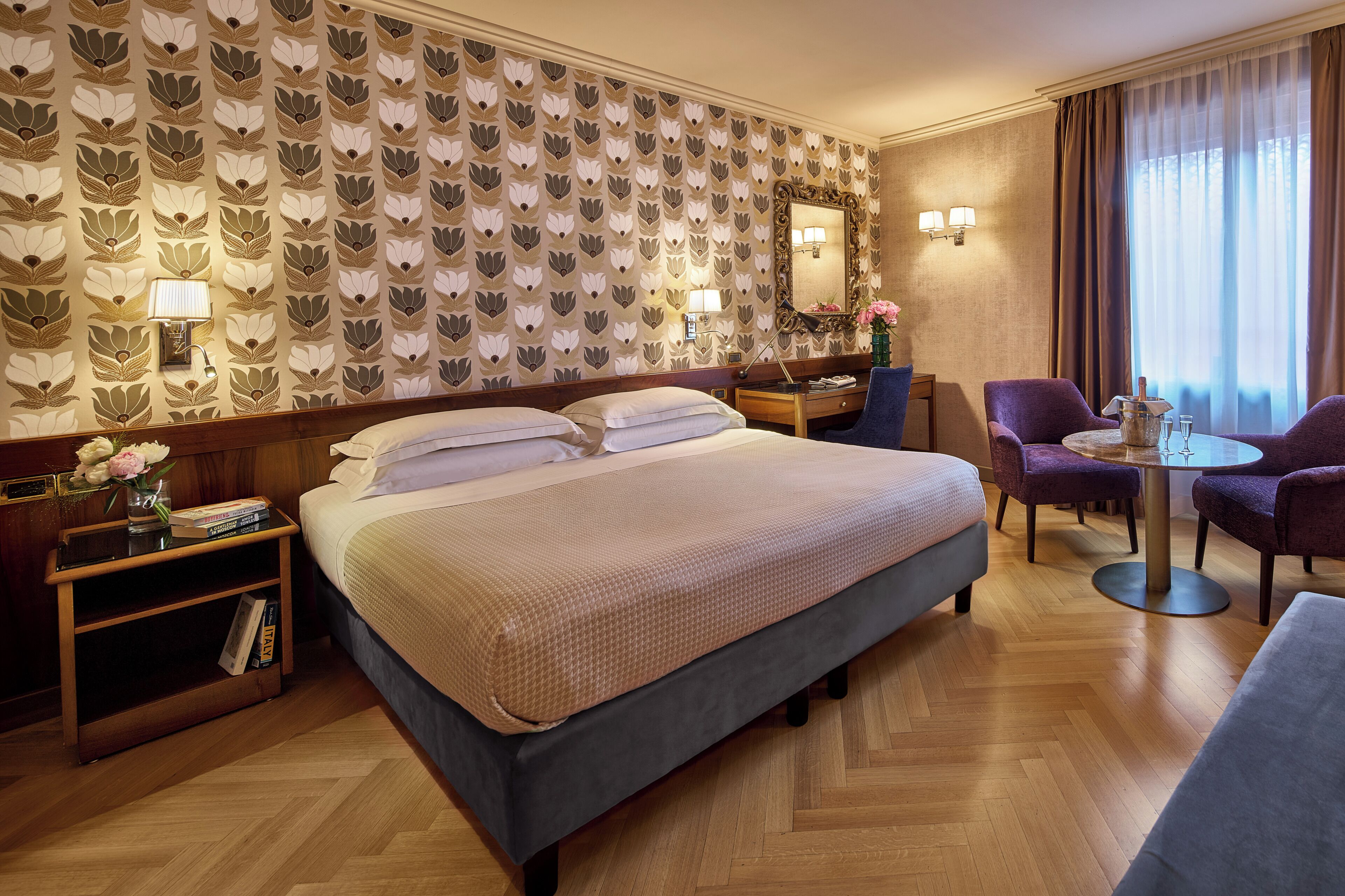 A hotel room with a large bed, a desk with a chair, a small table with chairs, and a decorative wall with a floral pattern.