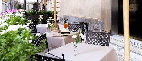 An outdoor dining area with tables covered by umbrellas, surrounded by greenery and flowers.