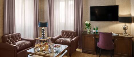 A well-appointed hotel room with leather armchairs, a glass coffee table, a desk with a computer, and a flat-screen TV mounted on the wall.