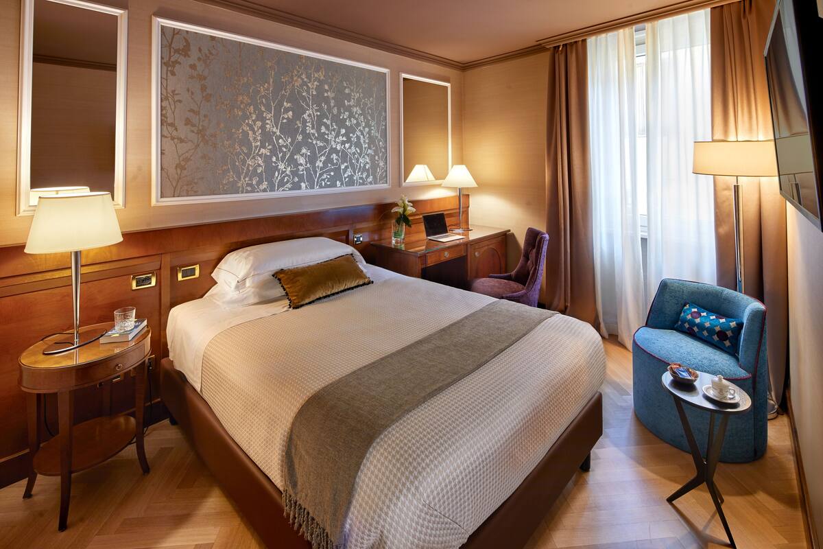 Une chambre d’hôtel avec un grand lit, un bureau avec une lampe, une chaise et une petite table sur laquelle se trouvent une tasse et une soucoupe.