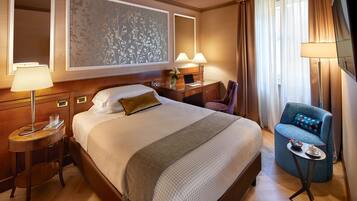 A hotel room with a large bed, a desk with a lamp, a chair, and a small table with a cup and saucer.