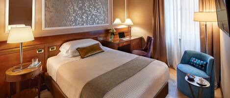 A hotel room with a large bed, a desk with a lamp, a chair, and a small table with a cup and saucer.