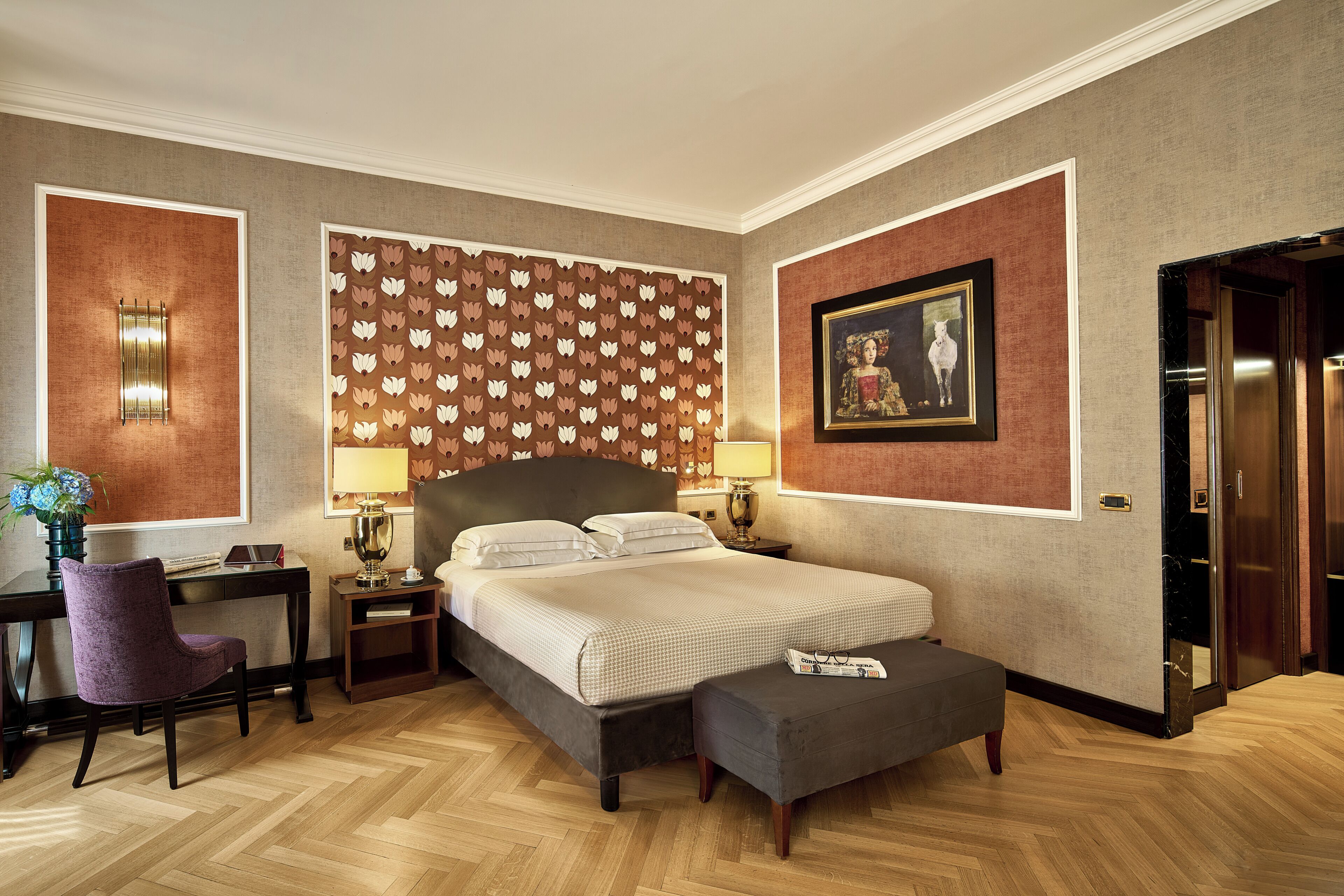 A hotel room with a large bed, a desk with a chair, a small table with a lamp, a wall-mounted picture, and a door leading to another room.