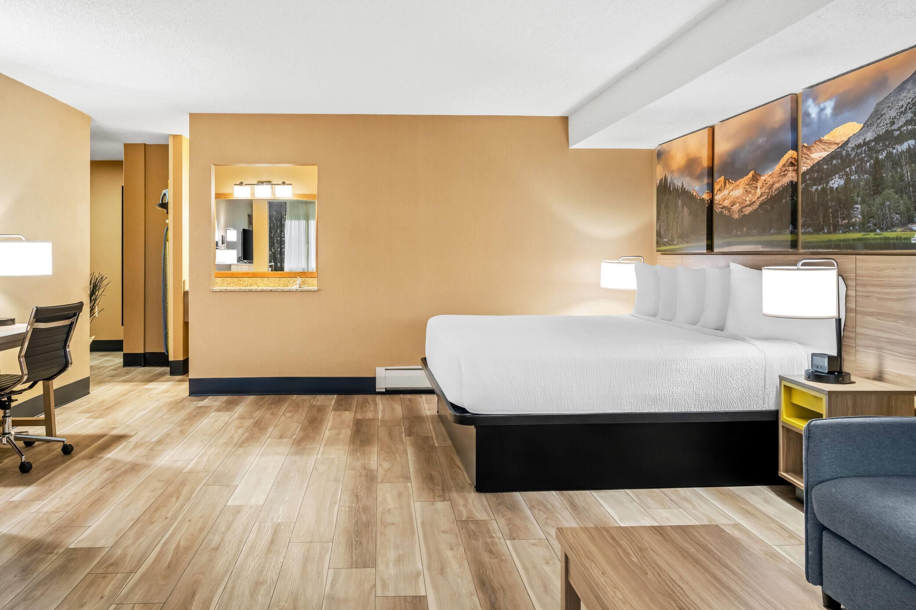 A hotel room with a bed, desk, chair, and a large painting of mountains.