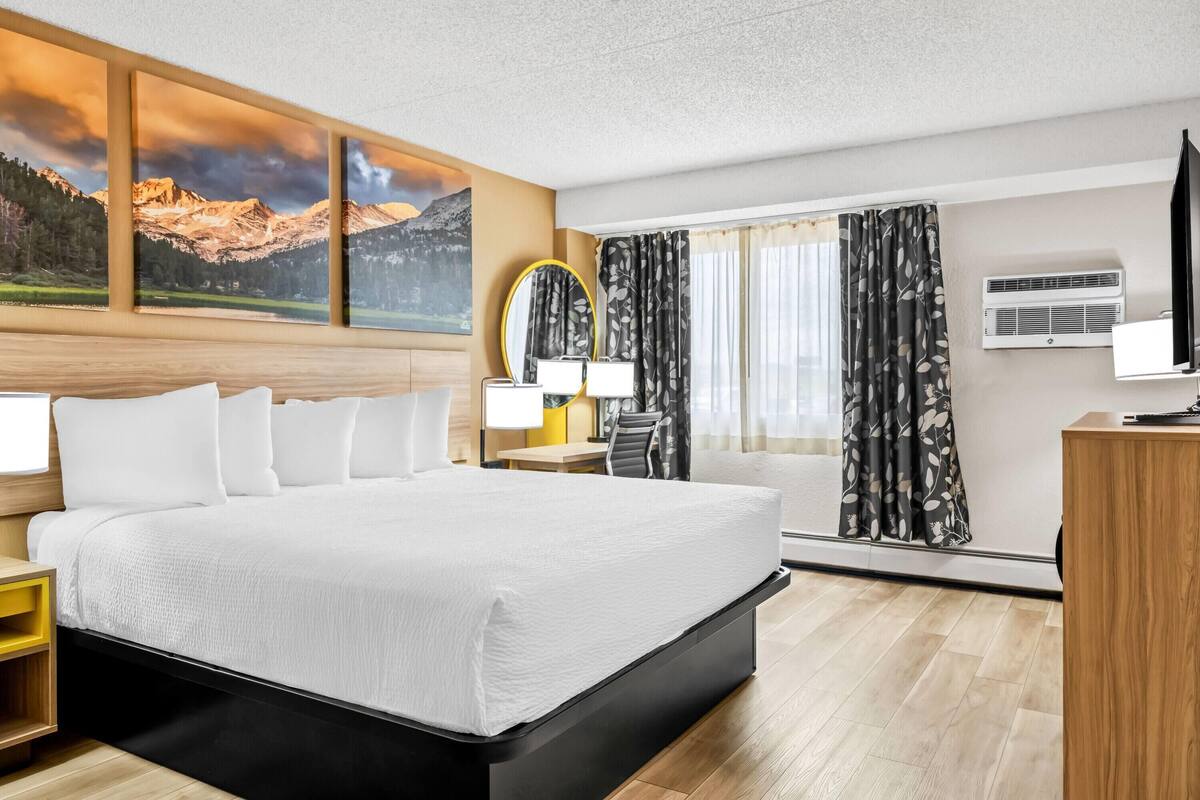 Ein Hotelzimmer mit einem großen Bett, einem Schreibtisch mit Lampe, einem Fernseher und einem Fenster mit Vorhängen.