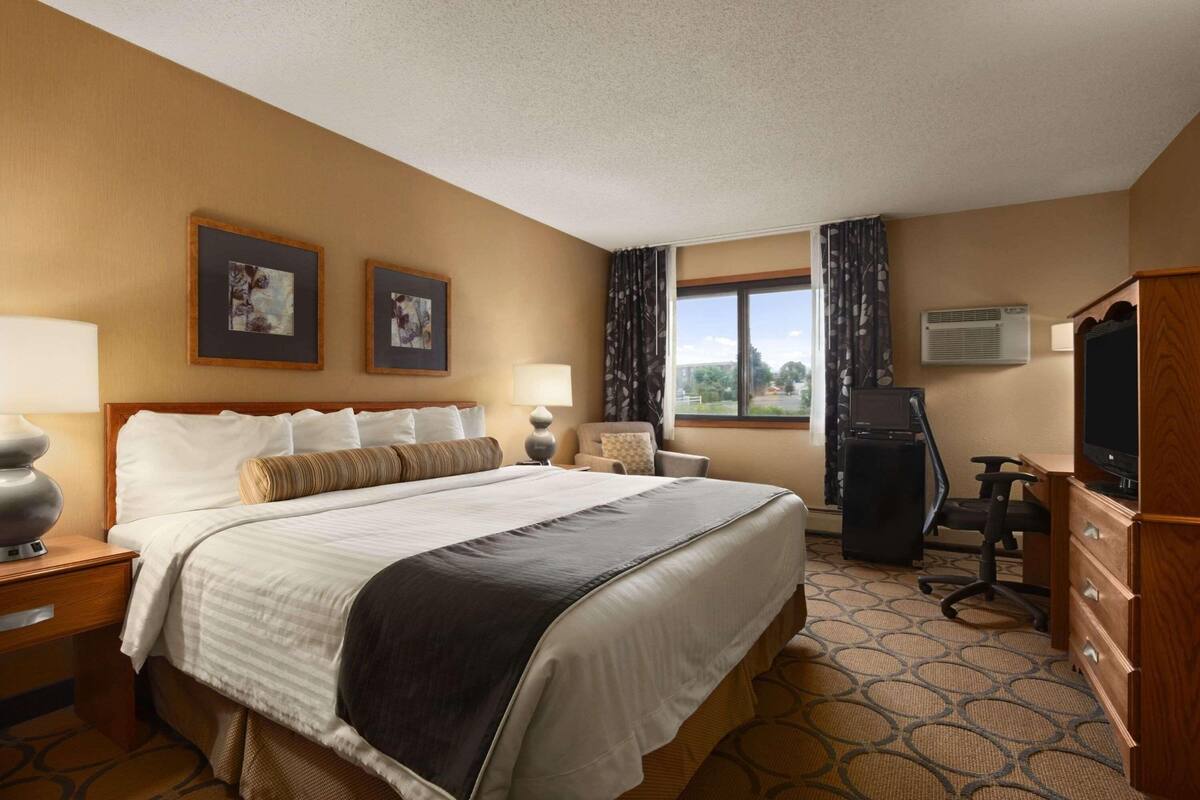 Ein Hotelzimmer mit einem großen Bett, einem Schreibtisch, einem Sessel, einem Fernseher und einem Fenster mit Vorhängen.