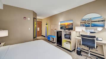 Ett hotellrum med en stor säng, ett skrivbord med en dator, en stol, en tv och utsikt över en solnedgång över vattnet.