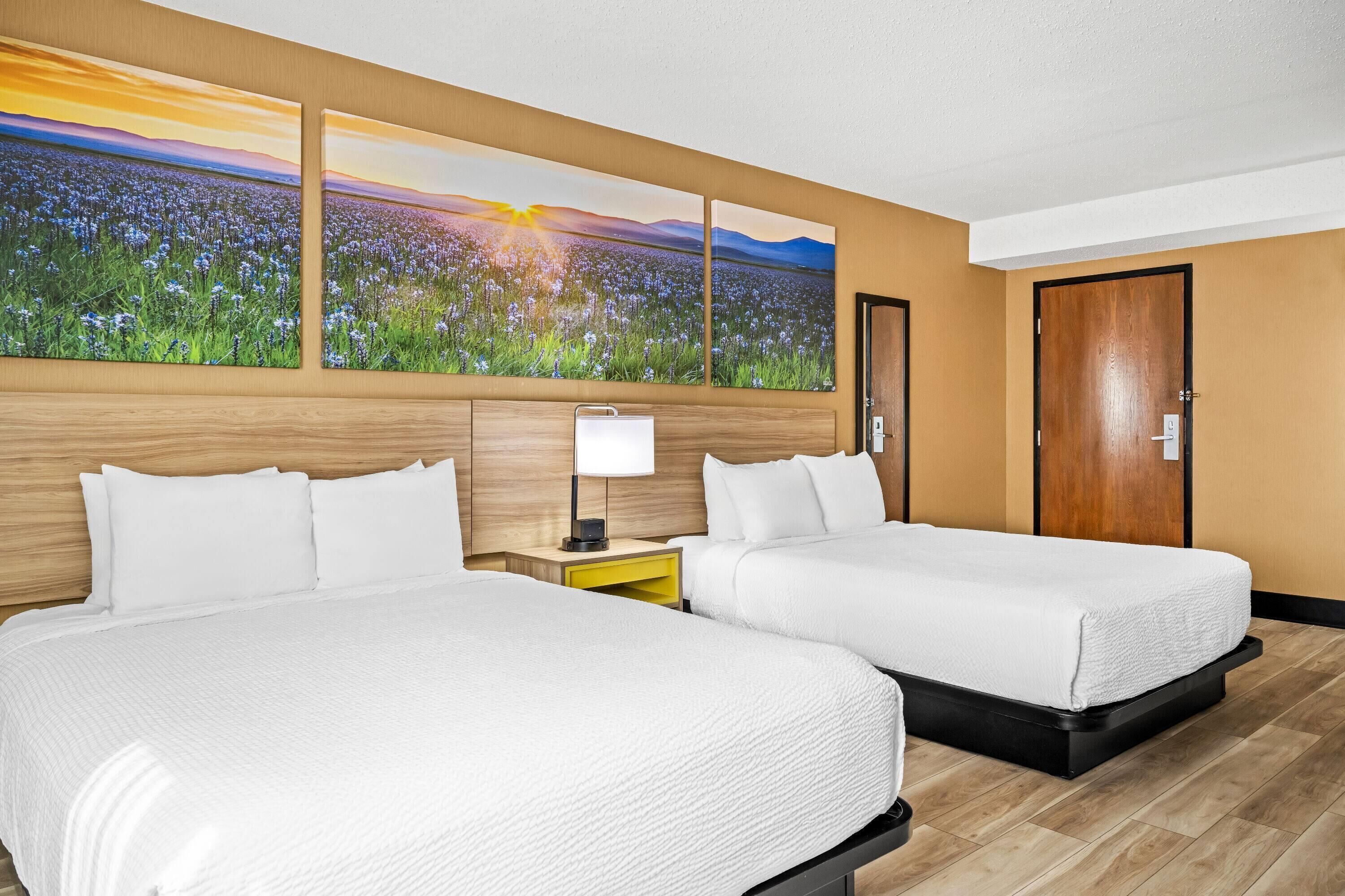 A hotel room with two beds, a wooden headboard, a nightstand with a lamp, and a large painting of a field at sunset.