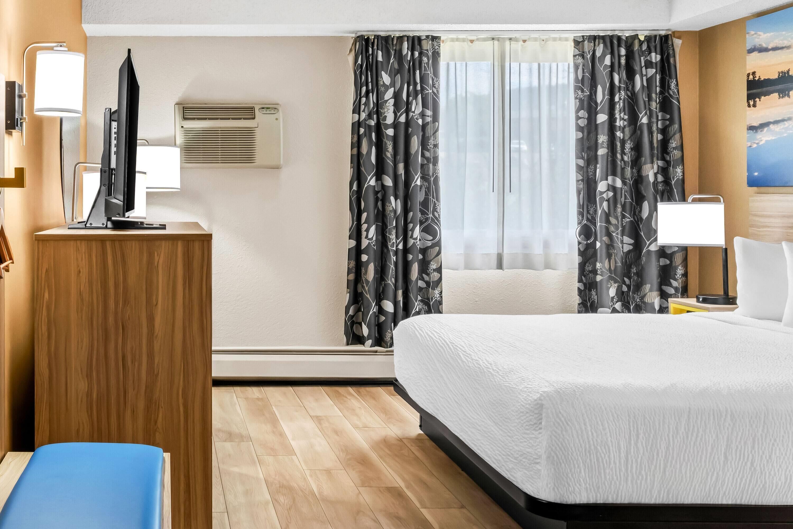Habitación de hotel con cama, escritorio con computadora, silla, lámpara y un cuadro en la pared.