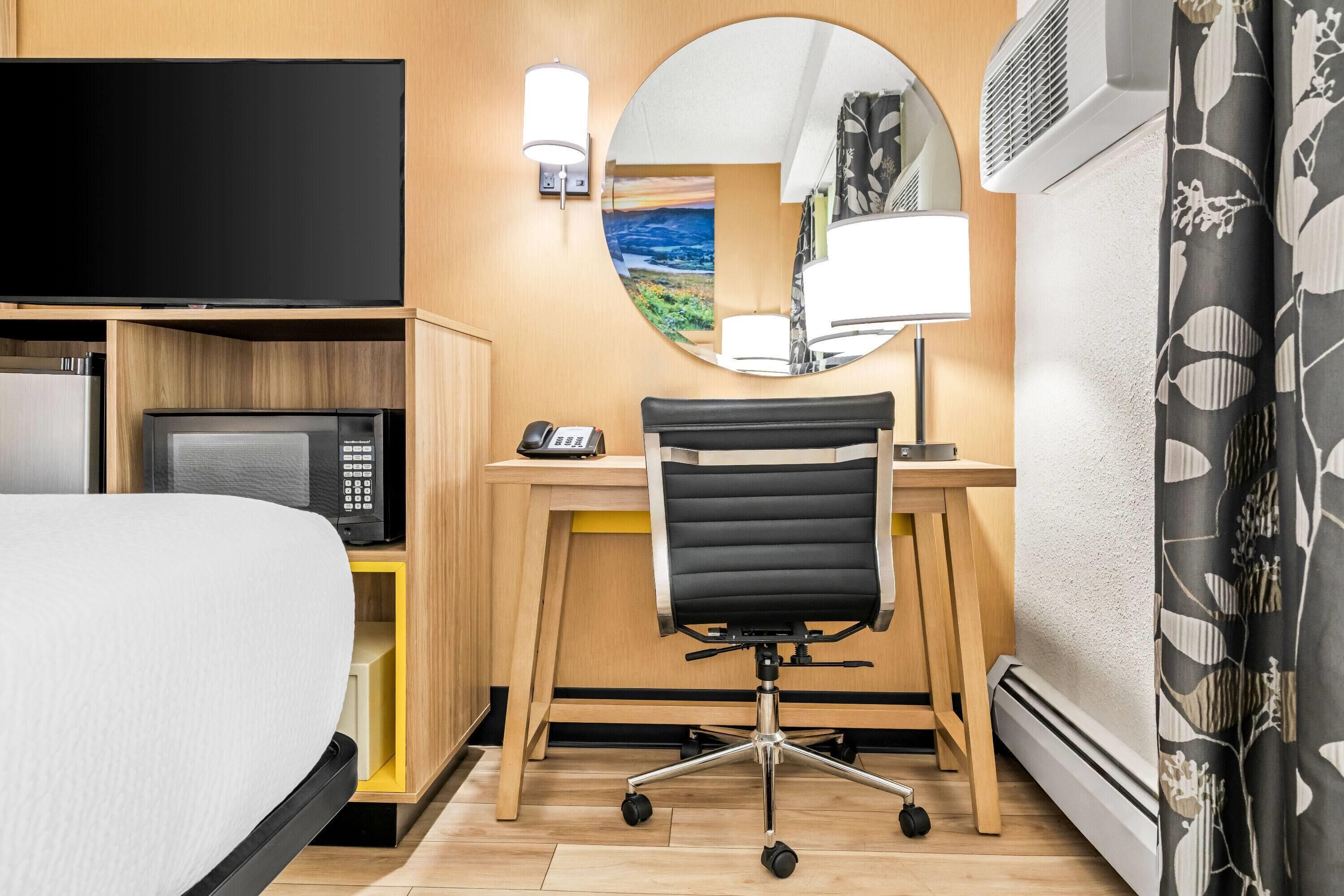 A hotel room with a bed, a desk with a computer, a chair, a TV, a lamp, and a mirror.