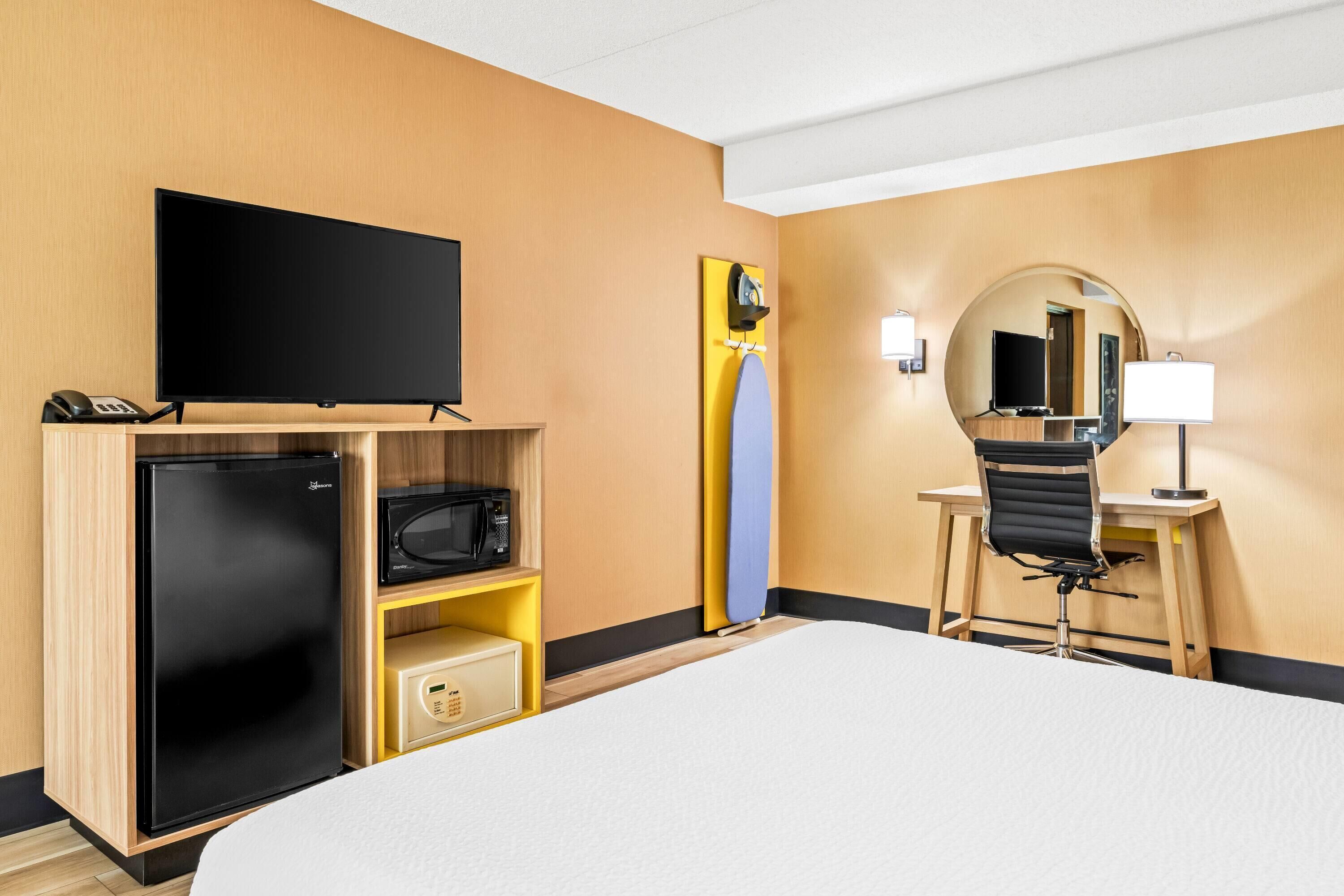 Camera d'albergo con un letto, una scrivania con una sedia, una televisione, un forno a microonde, un frigorifero e uno specchio.