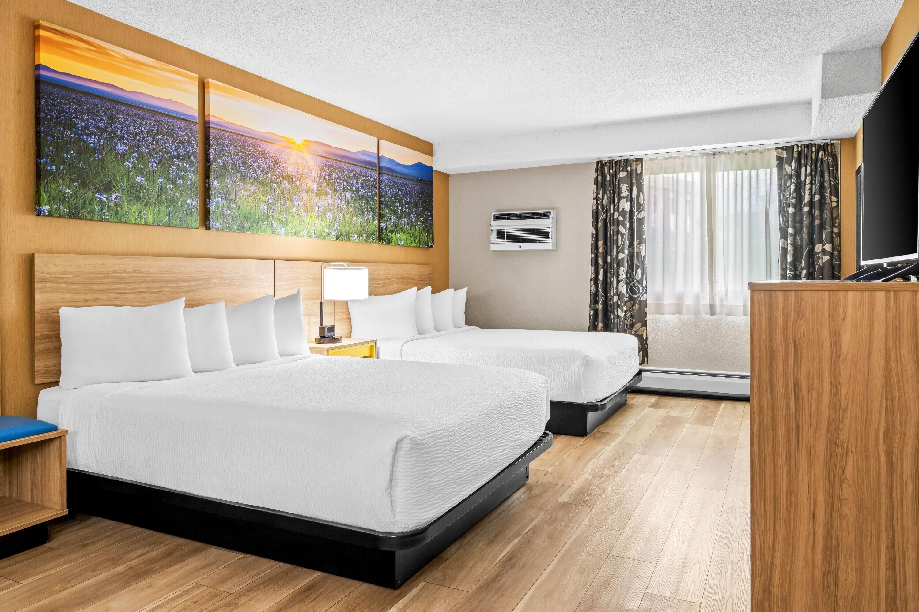 A hotel room with two beds, a large painting of a field, an air conditioning unit, a desk with a lamp, and a television.