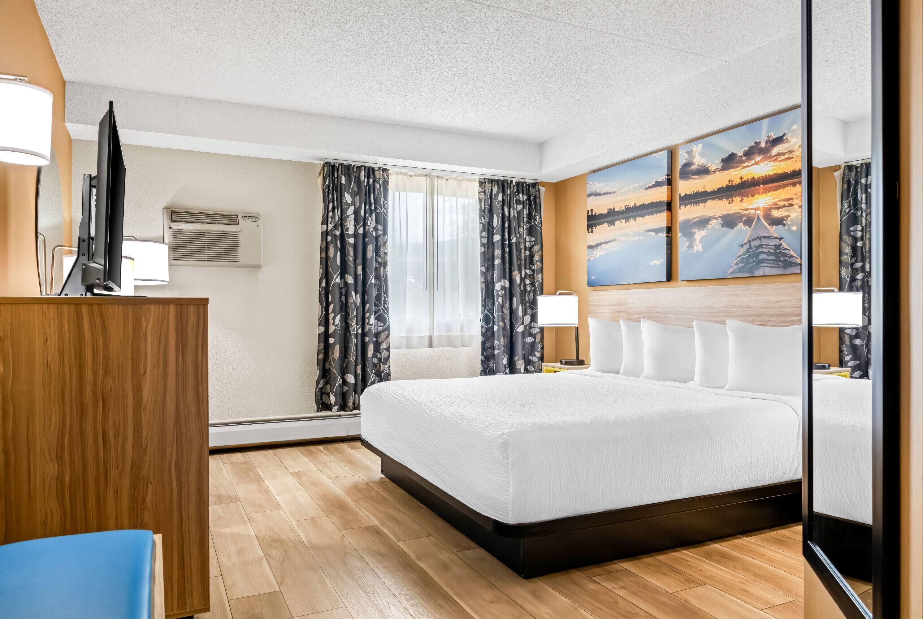Une chambre d’hôtel avec un grand lit, un bureau avec un ordinateur, une télévision et un tableau représentant un coucher de soleil sur un lac.