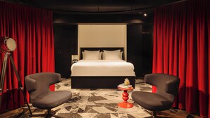 A hotel room with a large bed, red curtains, two armchairs, a small table with books, and a floor with a geometric pattern. - Leonardo Hotel Rotterdam Savoy (Rotterdam)