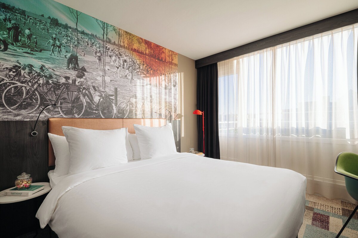 A hotel room with a large bed, a nightstand with a lamp, a chair, and a mural on the wall.