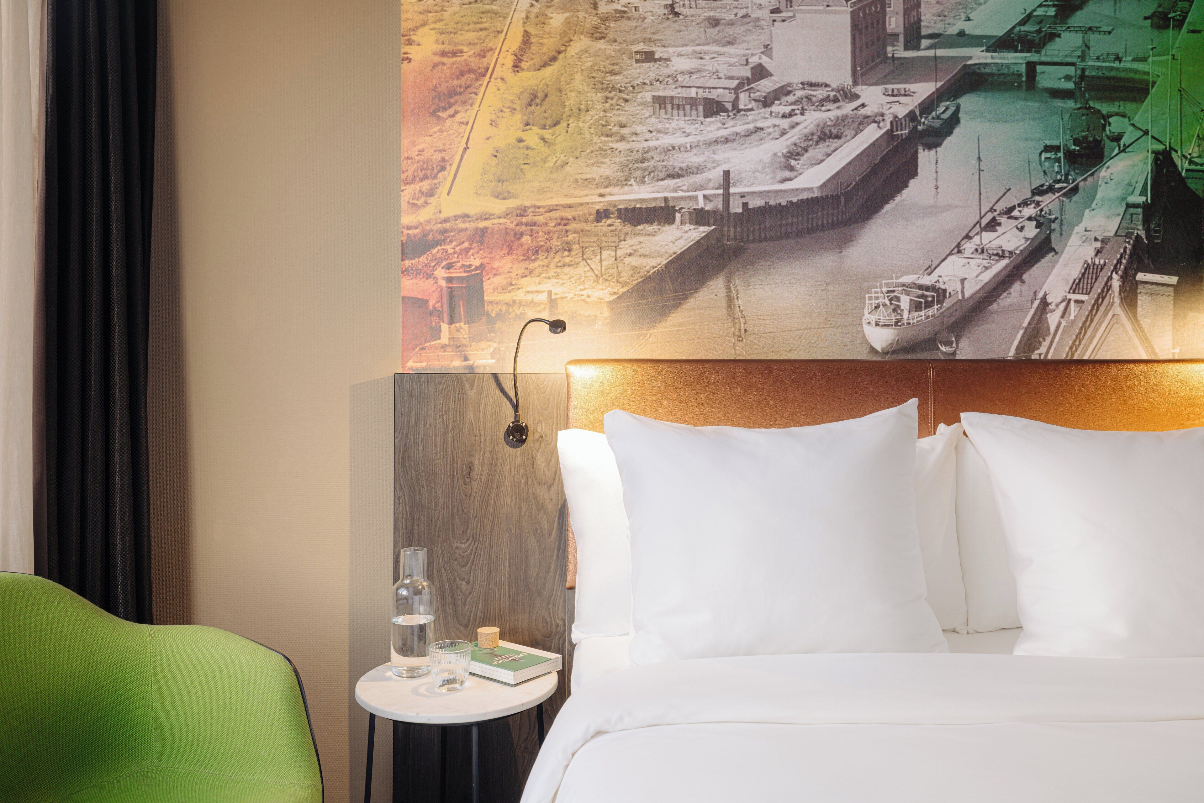 A hotel room with a bed, a green chair, a small table with a glass of water, a book, and a wall-mounted lamp.