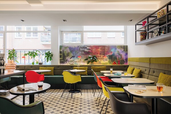 A modern cafe with a green sofa, colorful chairs, and a large window with a city view. - Leonardo Hotel Rotterdam Savoy (Rotterdam)