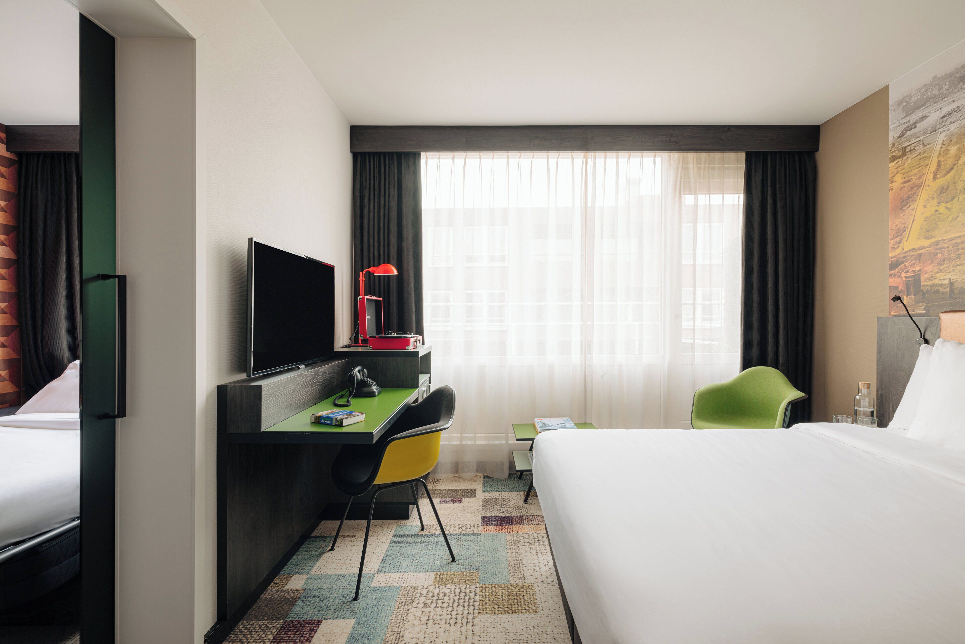 A hotel room with a bed, a desk with a computer, a chair, a TV, and a window with curtains.
