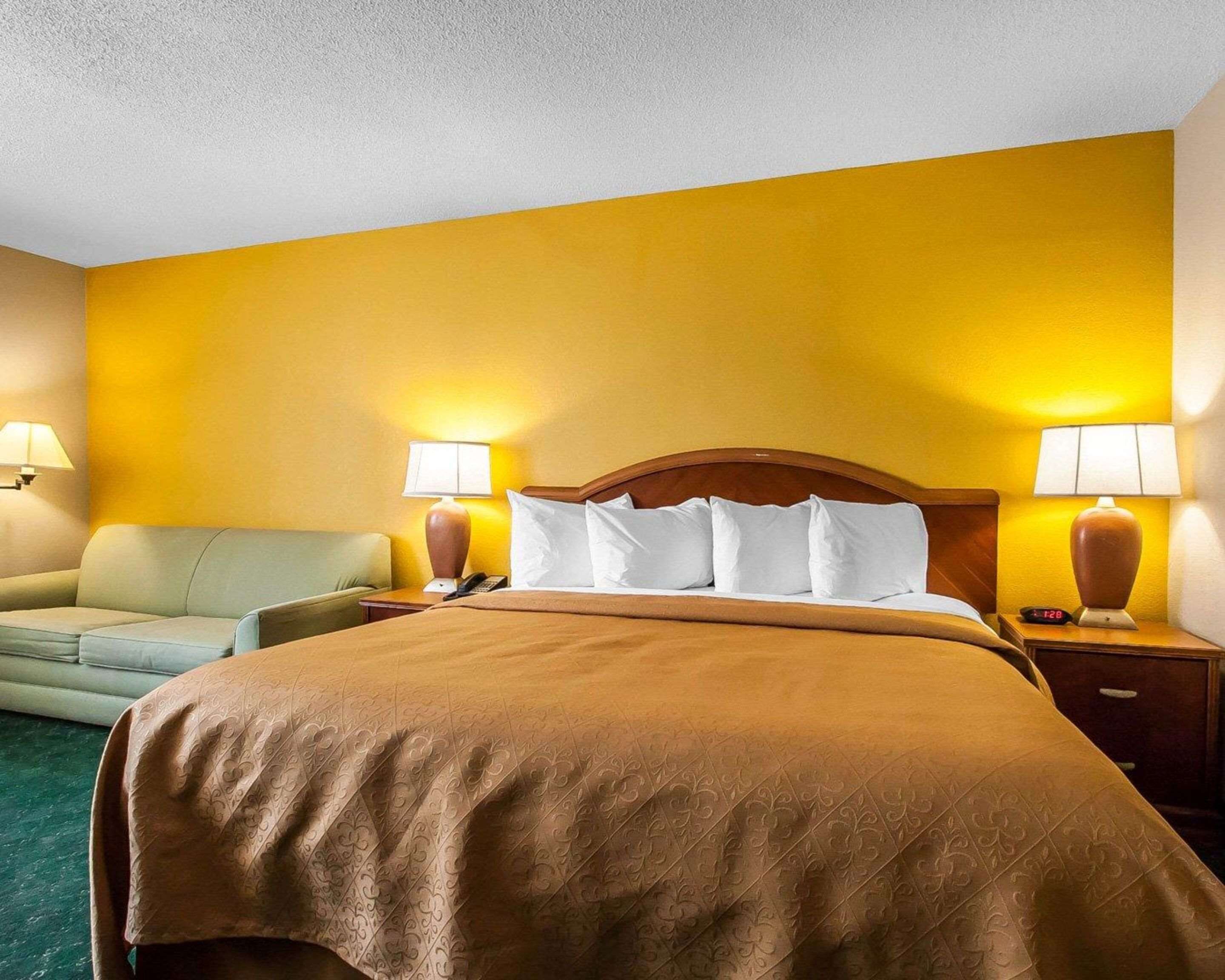 Quarto de hotel com uma cama grande, duas luminárias de cabeceira, uma mesa de cabeceira, um sofá e um abajur na parede.