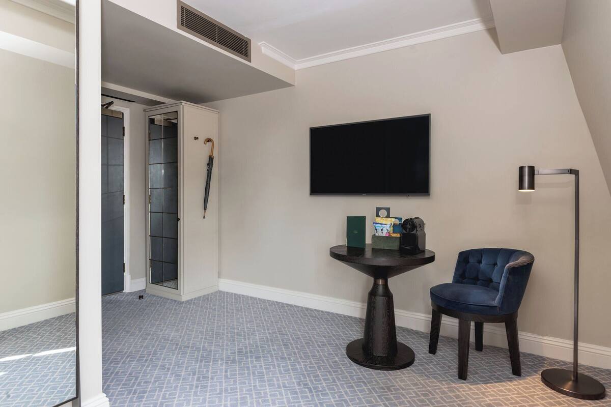 A room with a tiled floor, a flat-screen TV mounted on the wall, a small round table with a chair, and a floor lamp.