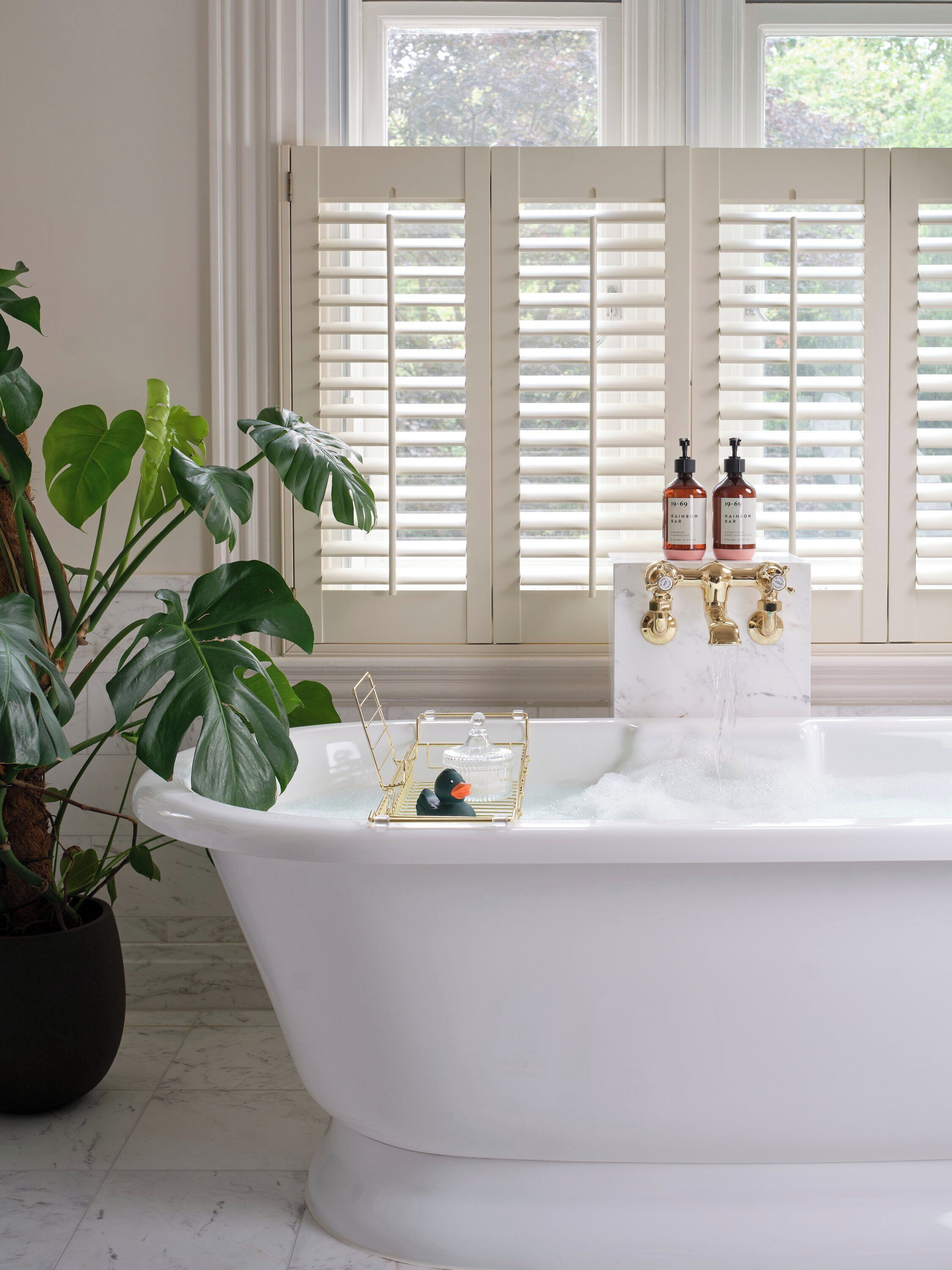 Une baignoire blanche avec un robinet doré, un distributeur de savon en forme de canard et un récipient en verre posé sur un plateau doré.