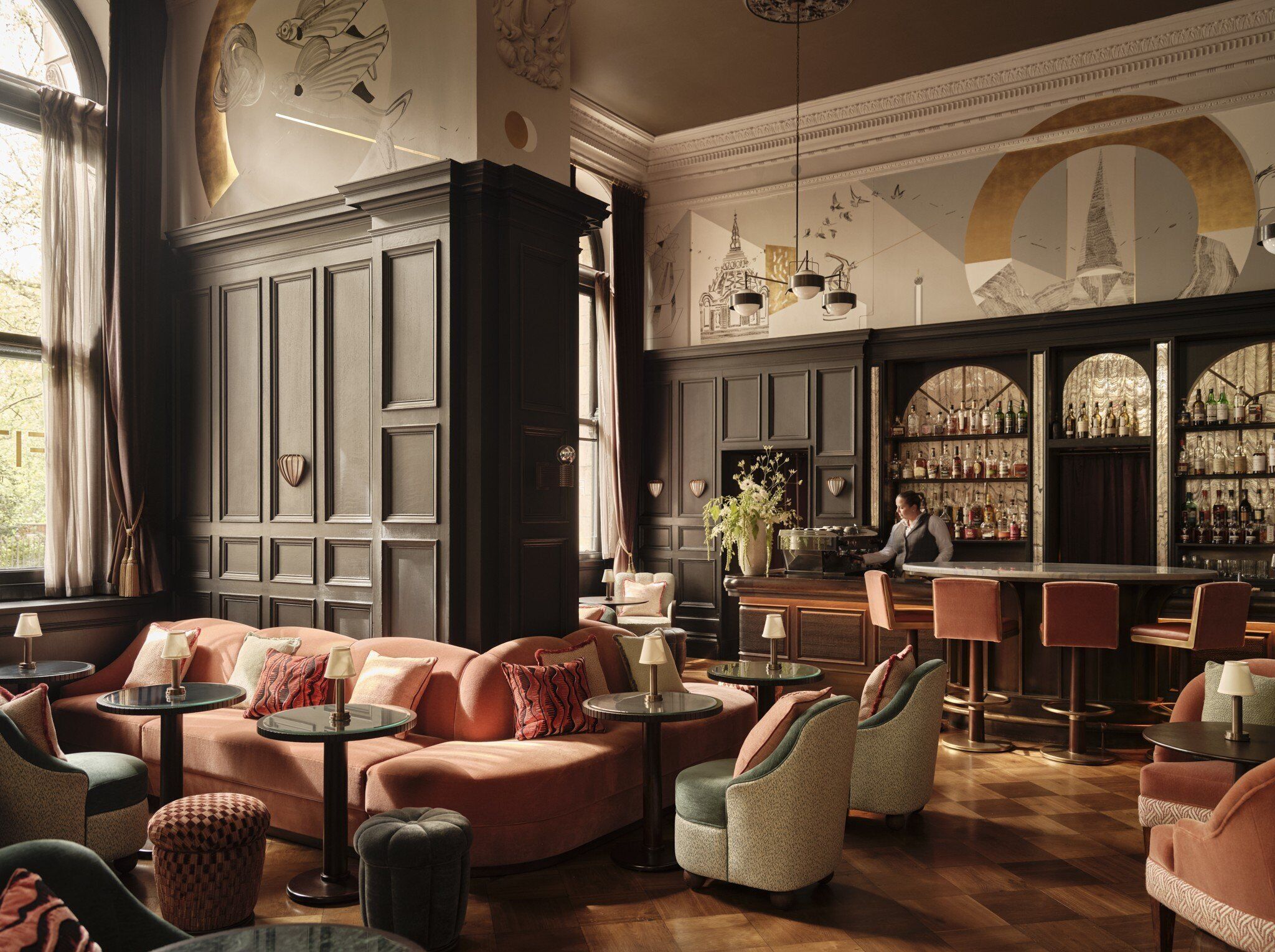 A bar area with a bartender, seating with round tables, and a wall with a mural.