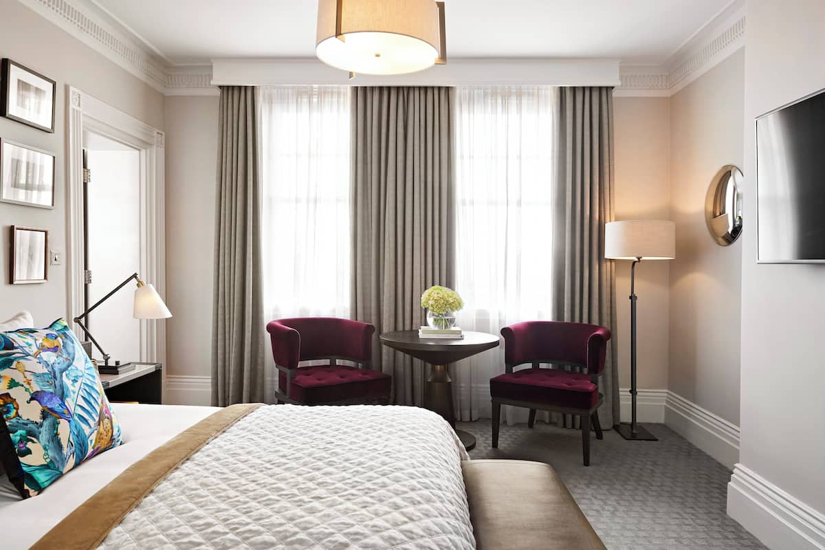 A hotel room with a bed, two chairs, a table with a vase, a lamp, and a TV on the wall.