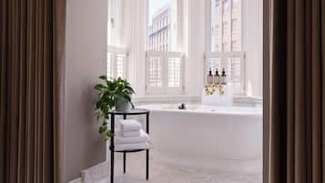 A room with a view of buildings through large windows, a plant on a table, and a bathtub with bottles.
