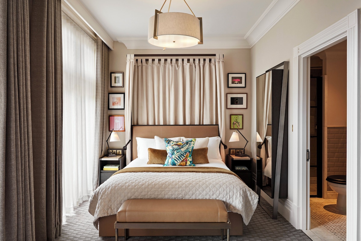 A hotel room with a large bed, bedside lamps, a bench, and a view of the bathroom.