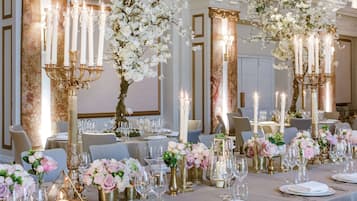 A formal dining room with tables set for a meal, featuring candelabras, floral arrangements, and elegant tableware.