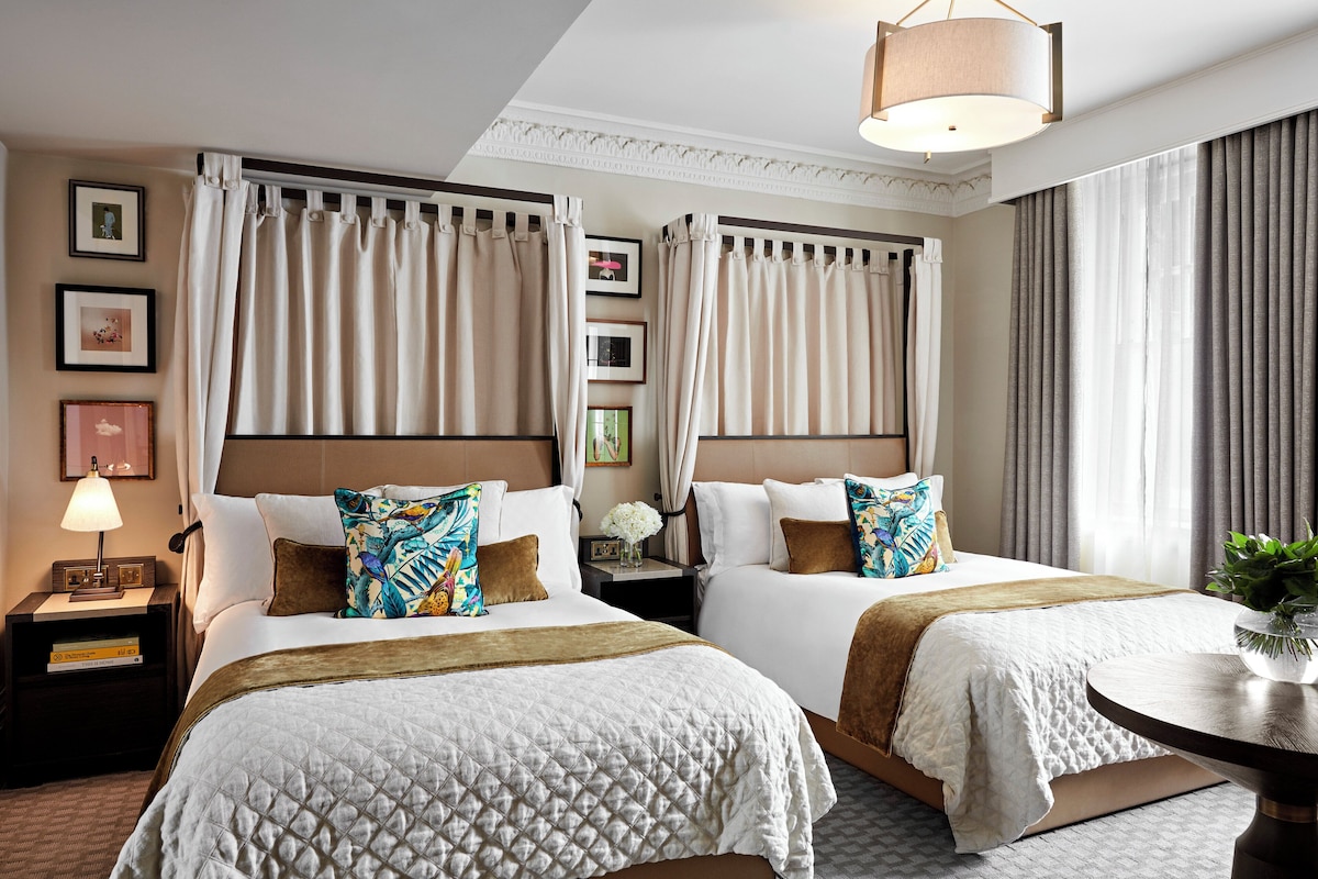 A hotel room with two beds, a bedside table with a lamp, a small table with a vase, and a view of the outside through the window.