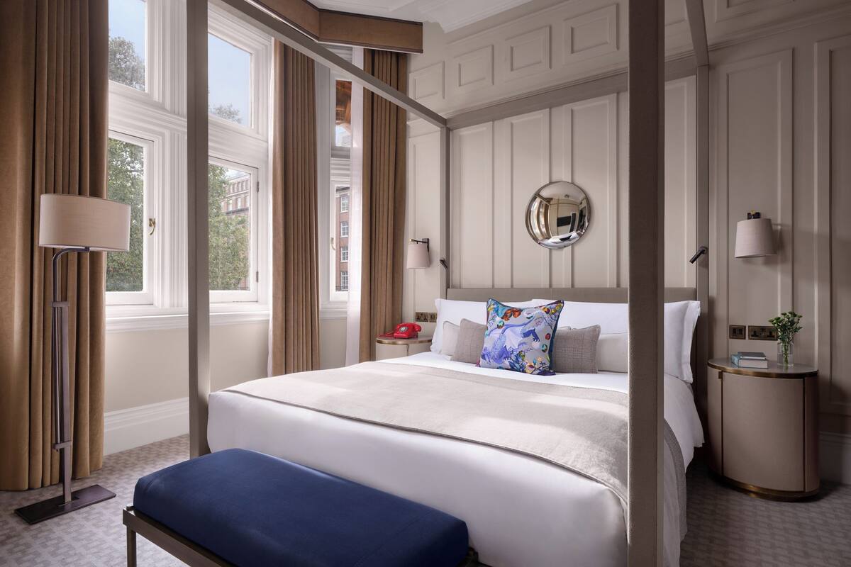 A hotel room with a large bed, a bedside table with a lamp, a round mirror on the wall, and a window with curtains.