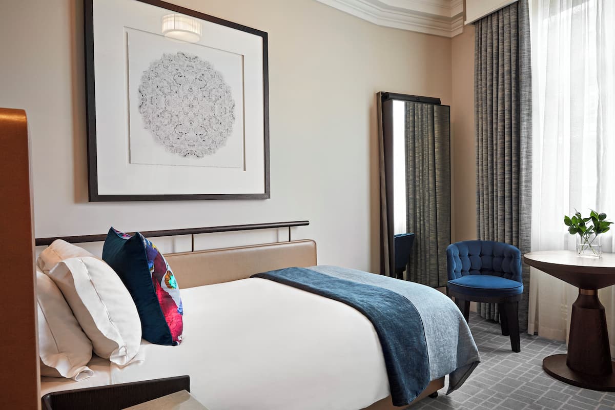 A hotel room with a bed, a chair, a table, and a framed artwork on the wall.