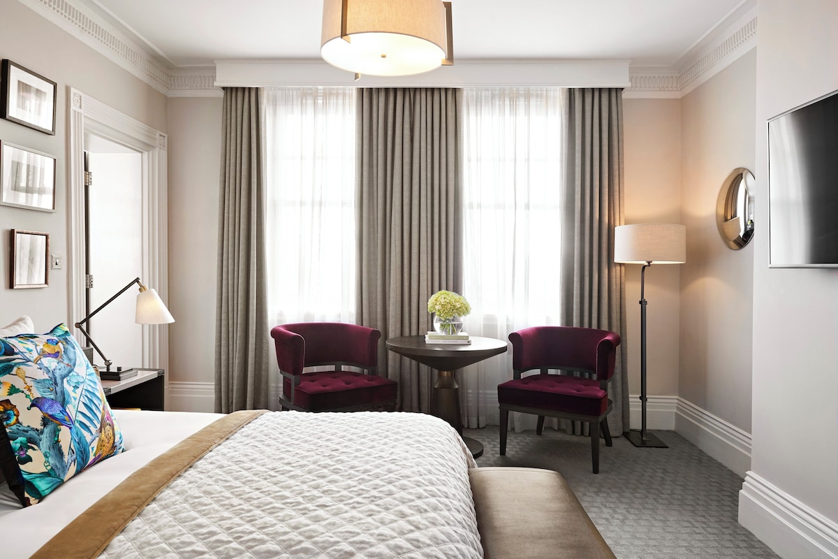 A hotel room with a bed, two armchairs, a small table, a lamp, and a TV.